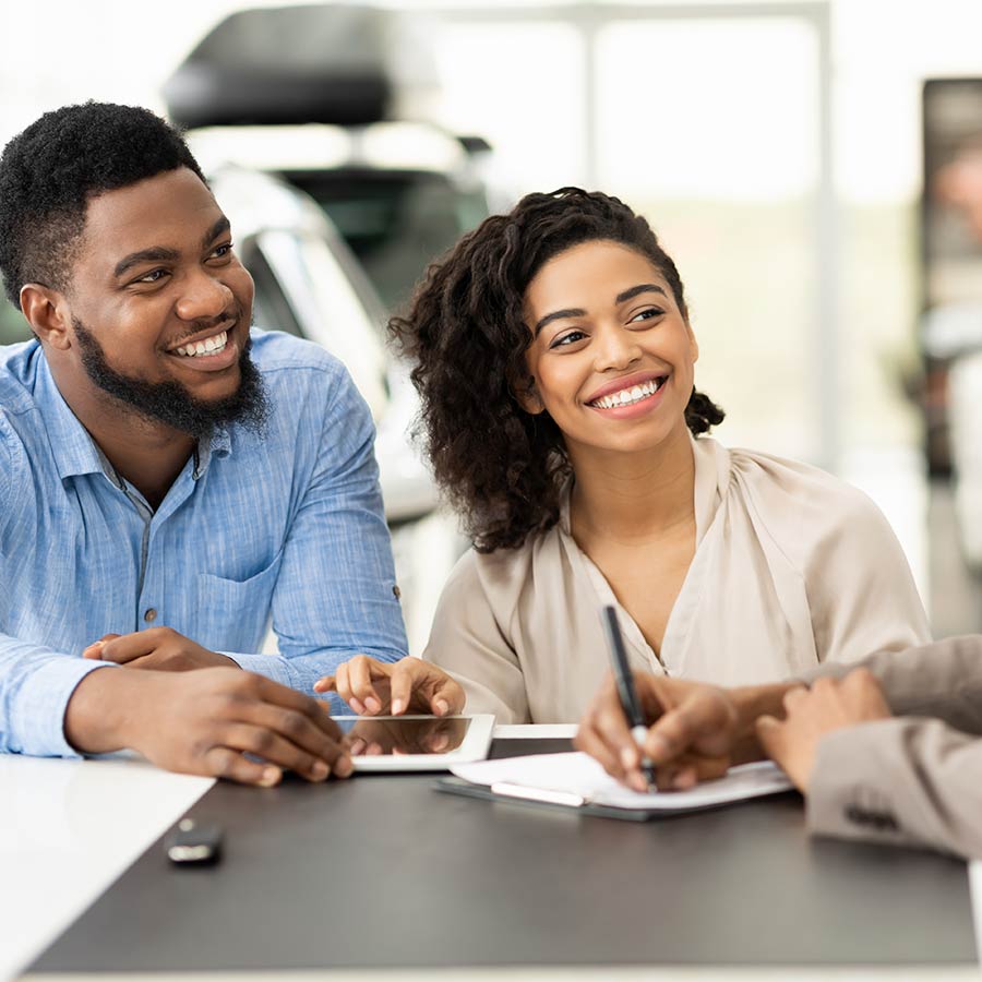 Couple Buying Car Signing Papers With Salesman In Dealership Office